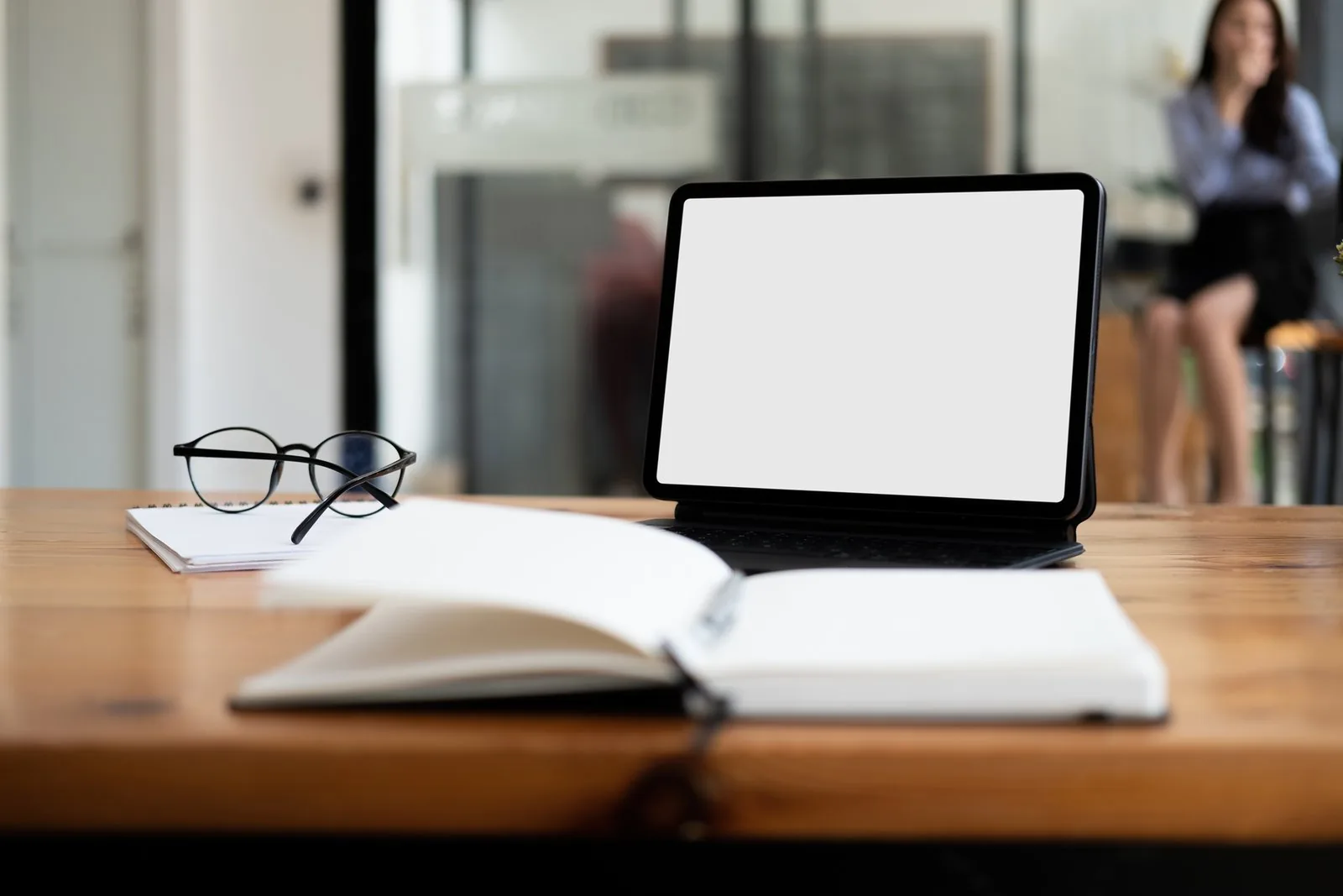 A tablet with a blank screen sits on a desk with notebooks, glasses, and a blurred person.