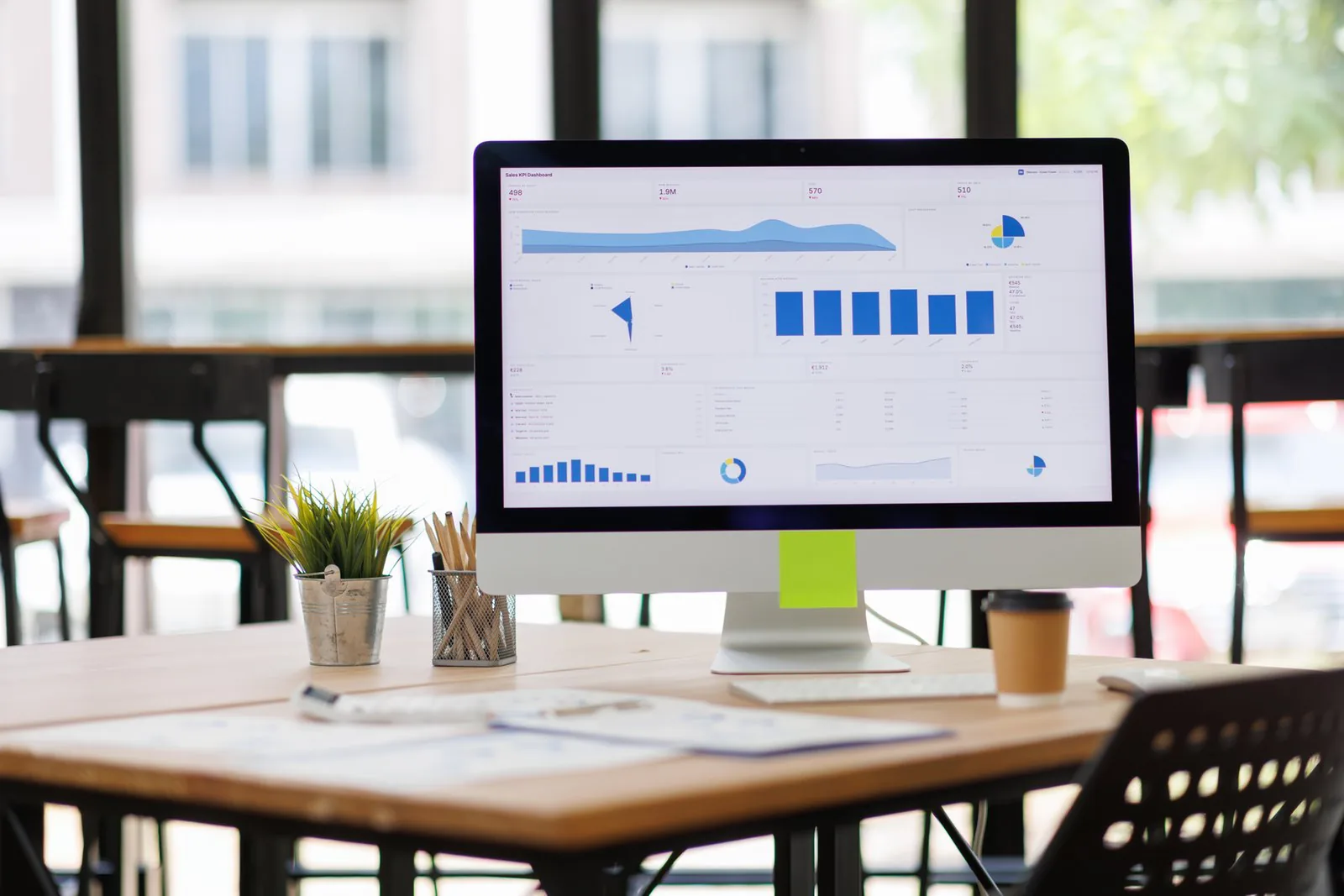 A desktop monitor shows blue charts on a wooden table with a plant, pencils, papers, coffee cup, keyboard, and windows.