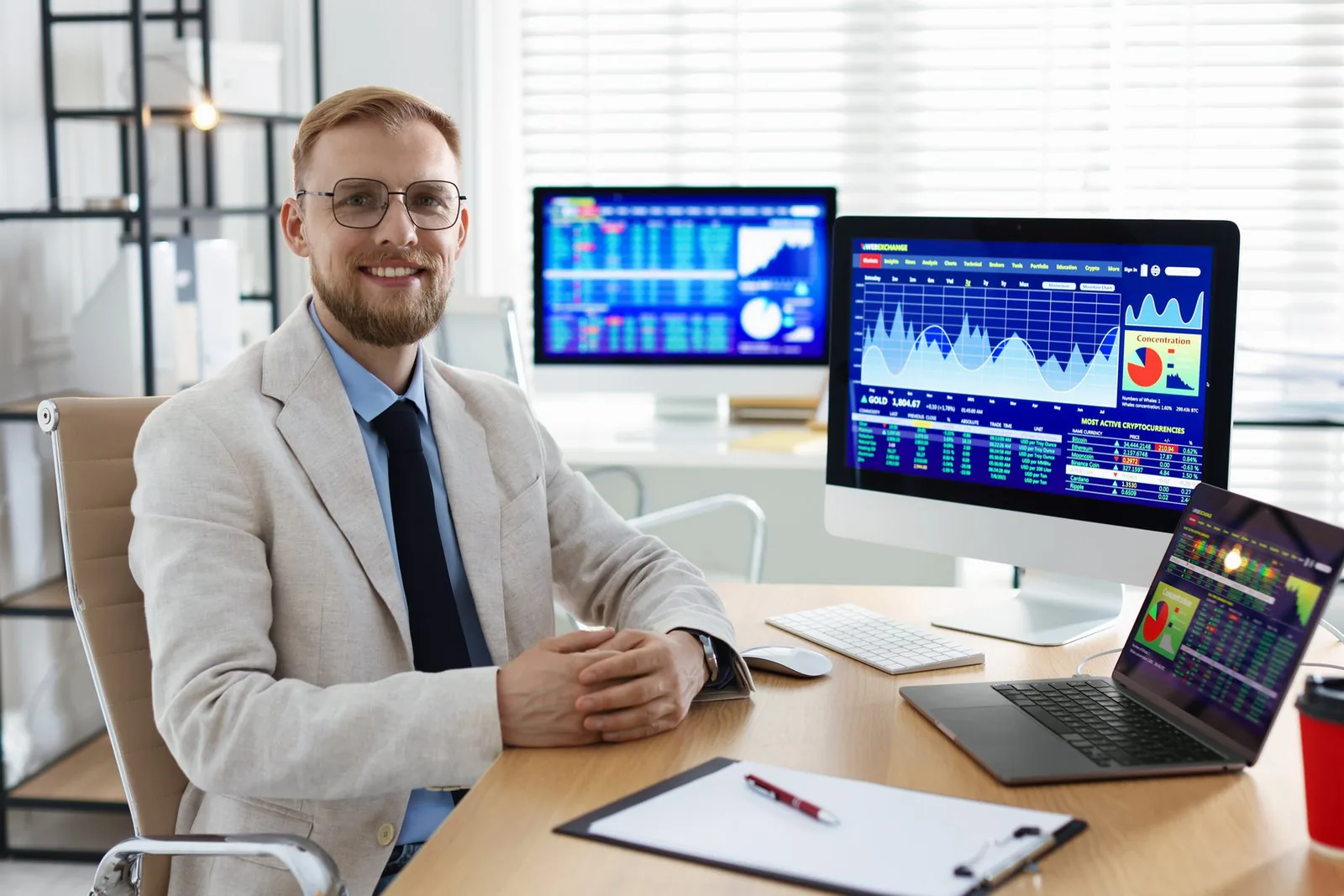 Smiling person in glasses sits at a desk with multiple screens showing colorful charts.