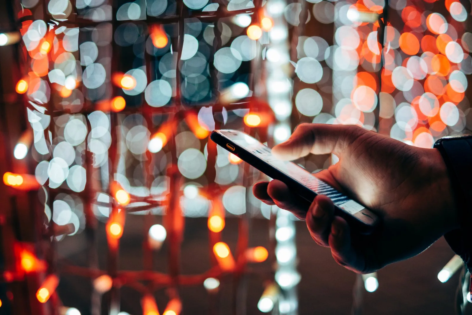 A hand uses a smartphone beside red and white string lights with a blurry background.