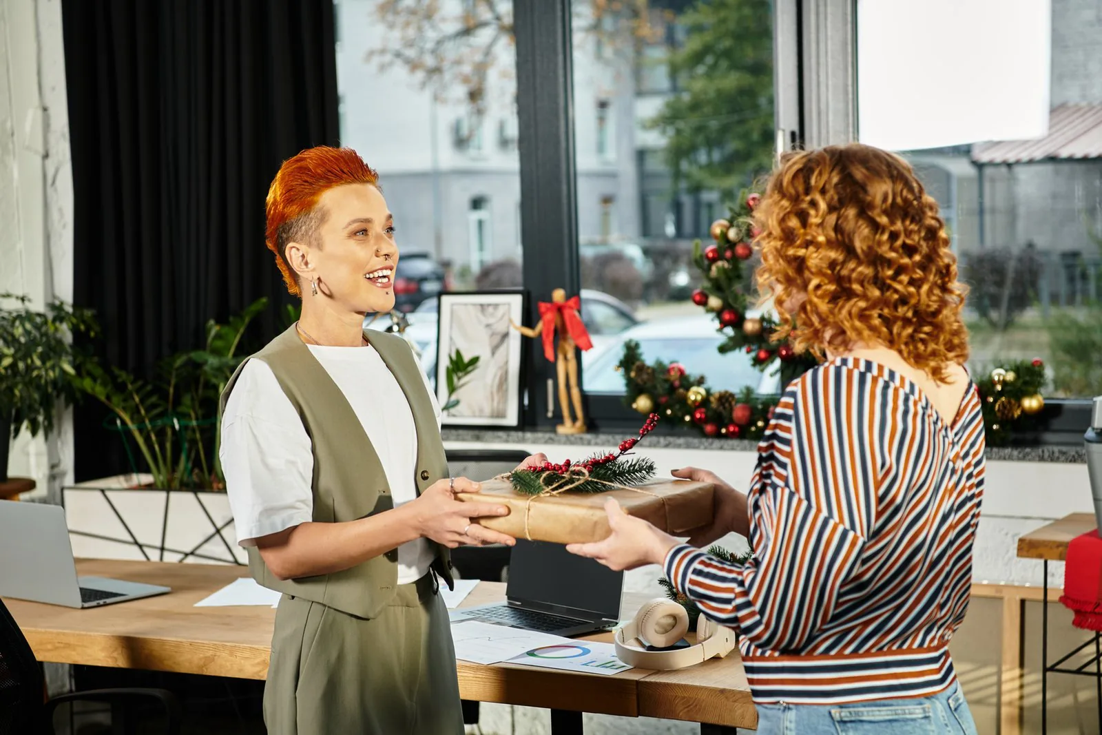 Two people in an office exchange a wrapped gift over a desk, with laptops, a wreath, and plants by the window.