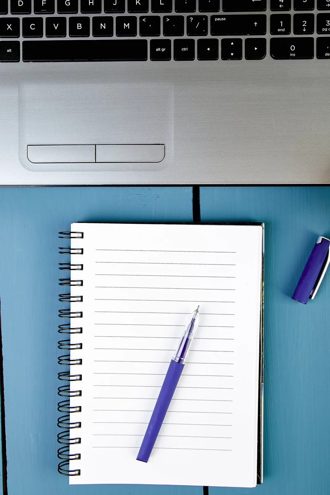 A laptop keyboard and touchpad, lined spiral notebook with purple pen, and pen cap on a blue table.