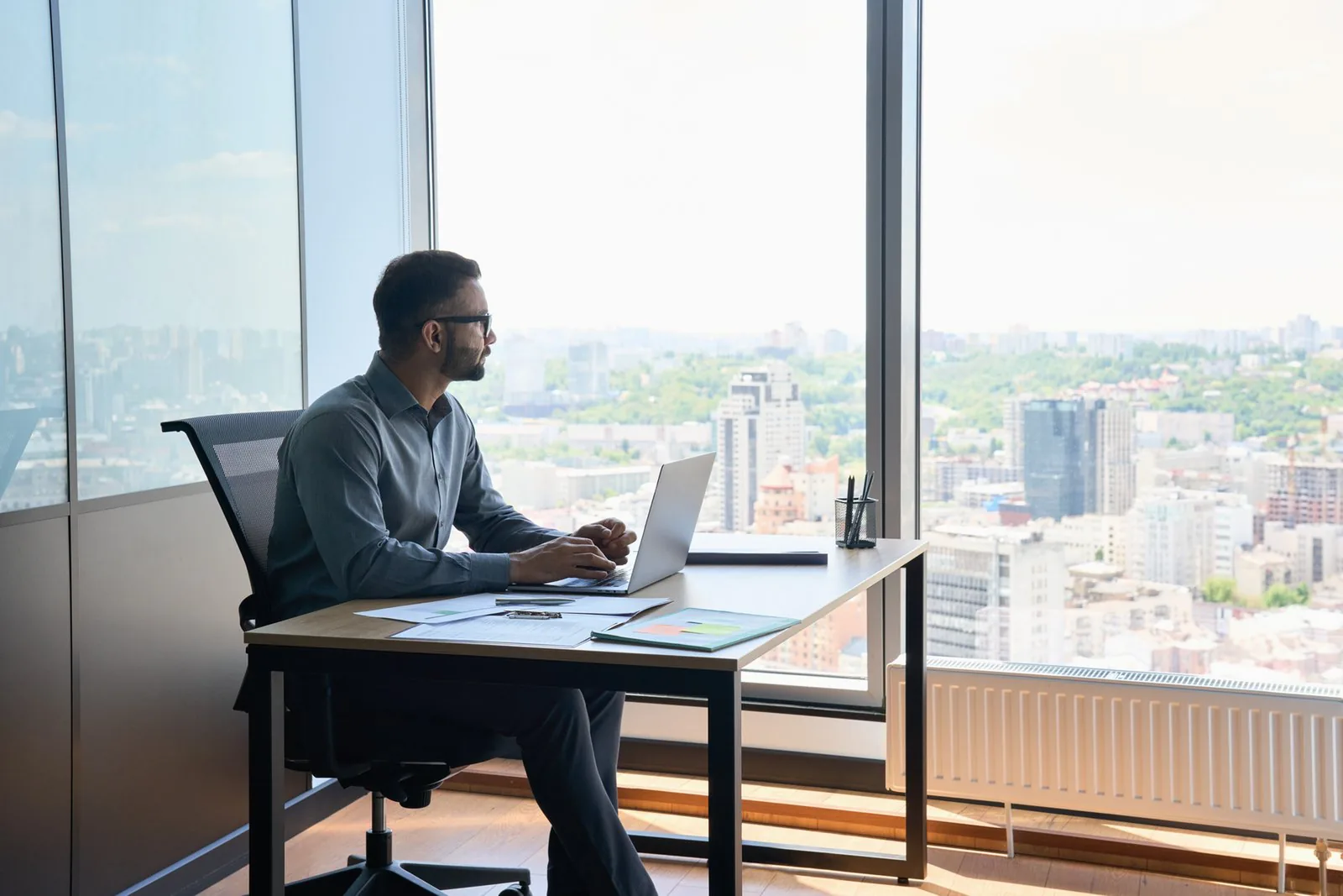 Man working at a laptop on a desk by tall windows overlooking a city.