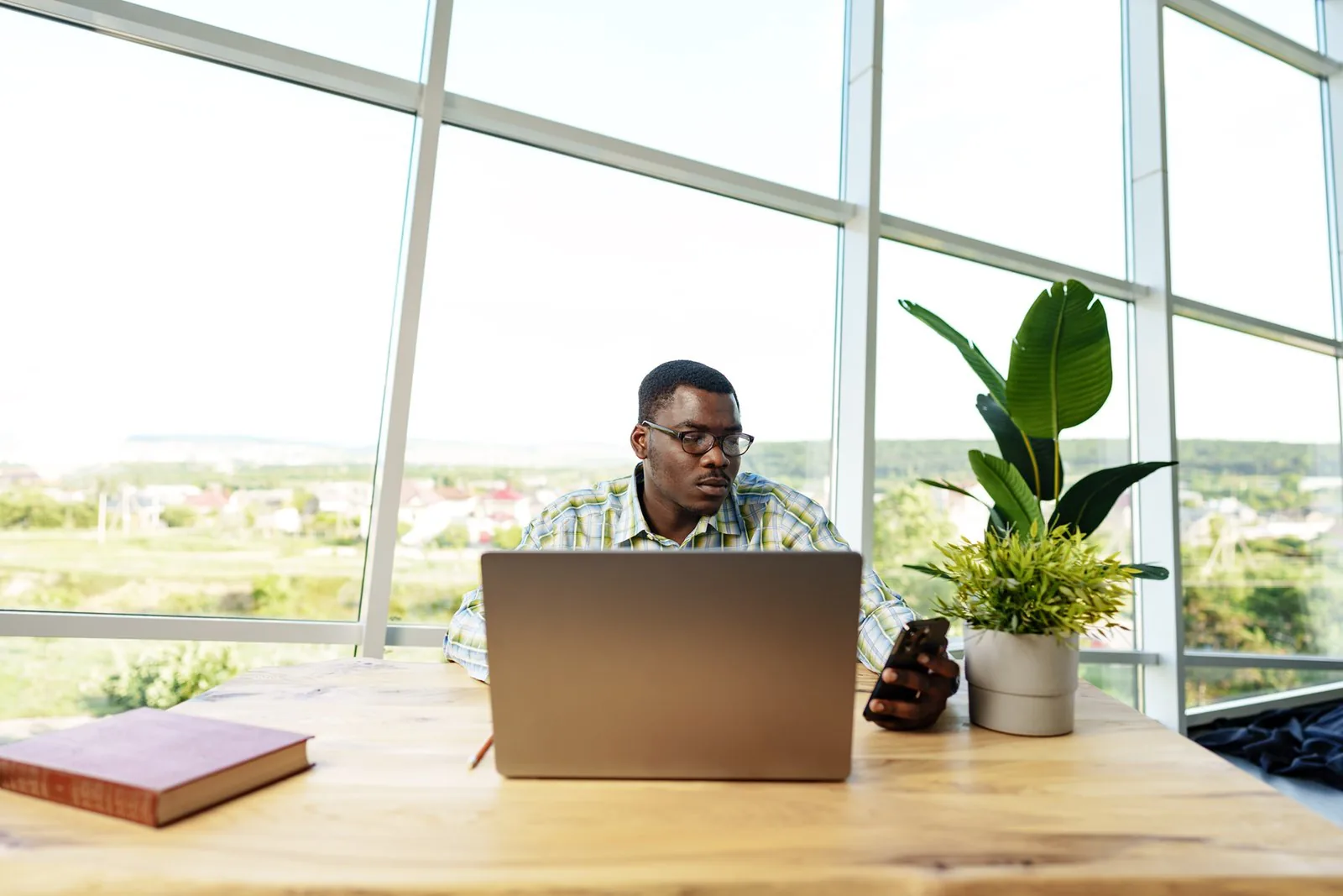 Person in glasses at a wooden table checks a phone beside a laptop, with a plant, book, and large windows.