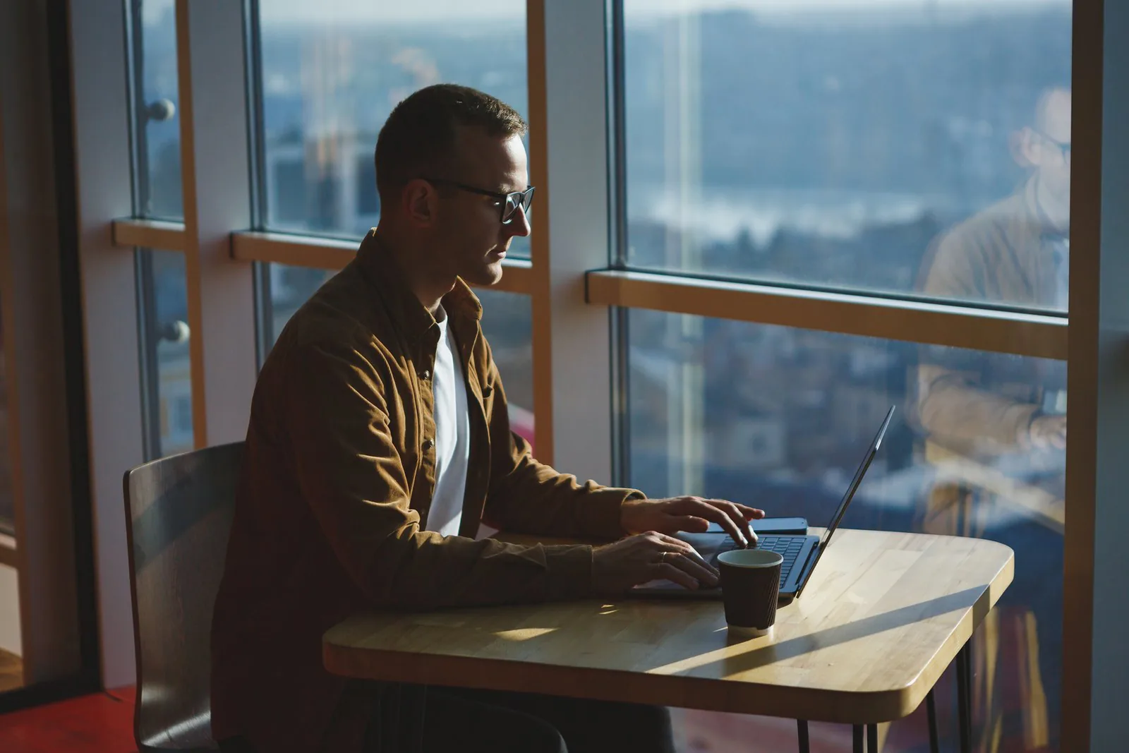 A person wearing glasses types on a laptop beside large windows, with a coffee cup and city view.