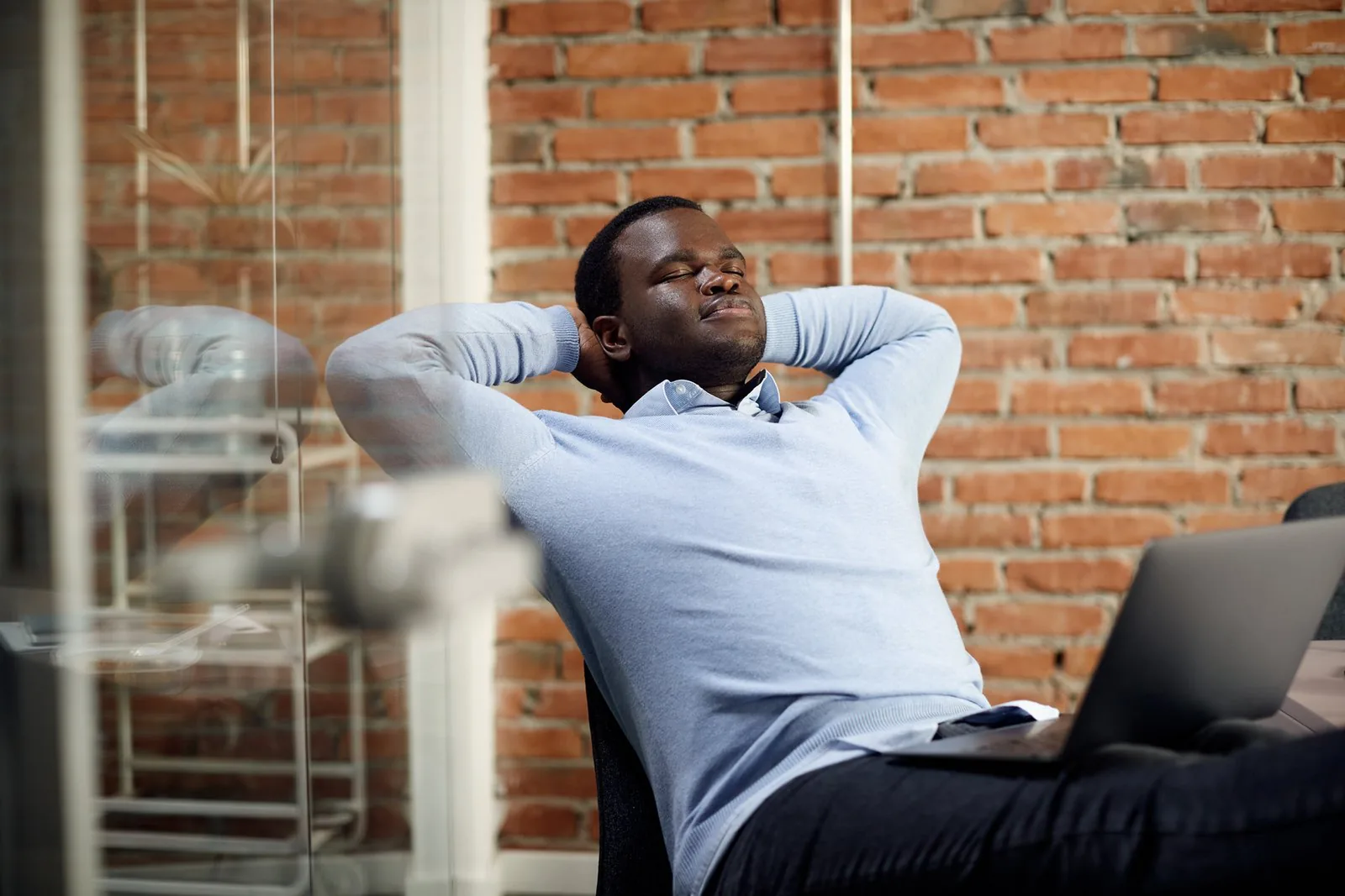 A person leans back in an office chair, eyes closed, hands behind head, with a laptop and brick wall.